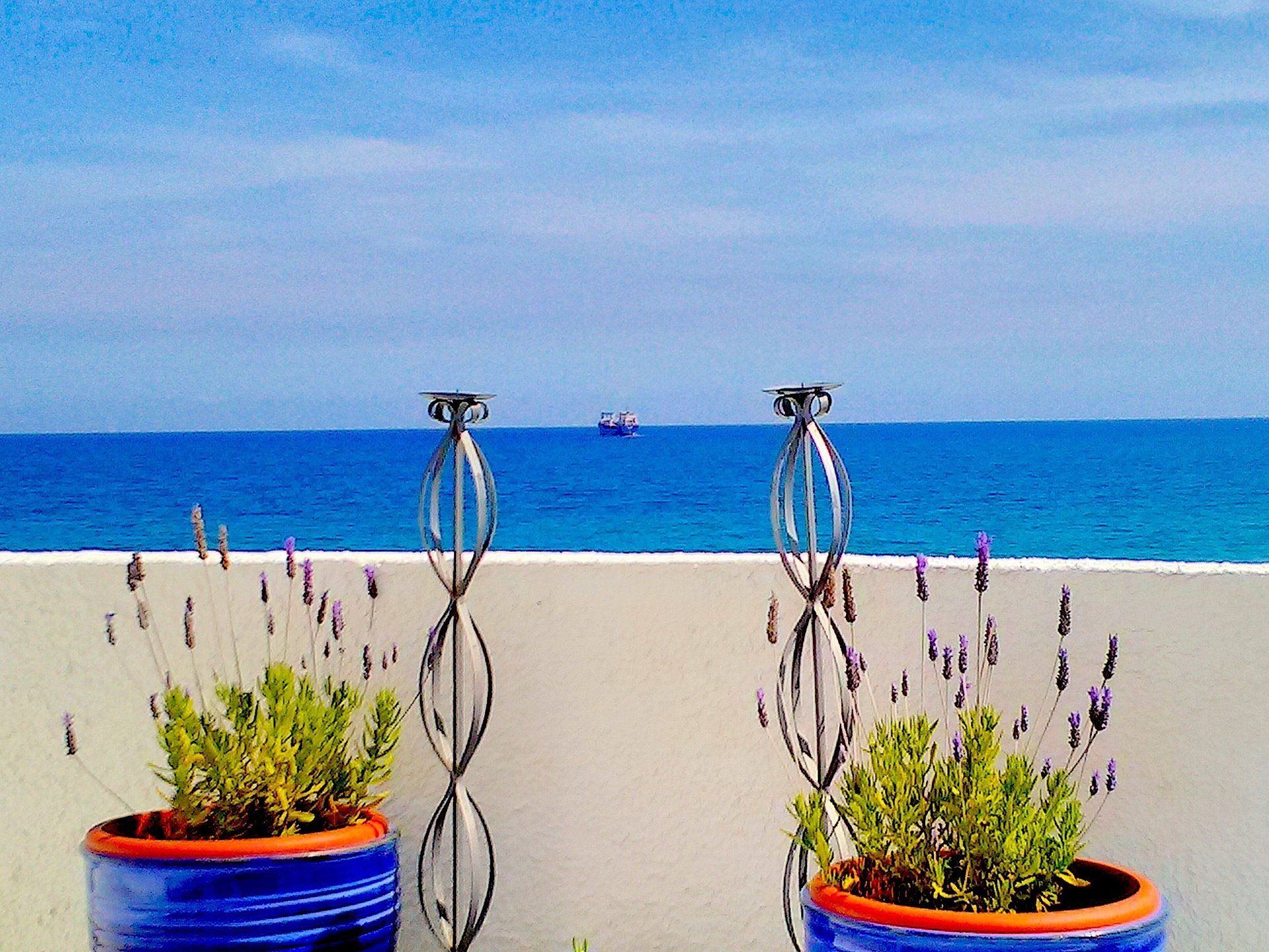 taken from a terrace two big pots of lavendar and two candlesticks frame a ship on the sea in the distance