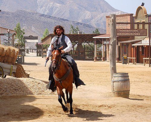 man dressed as cowboy on chestnut horse galloping towards you with western set in background