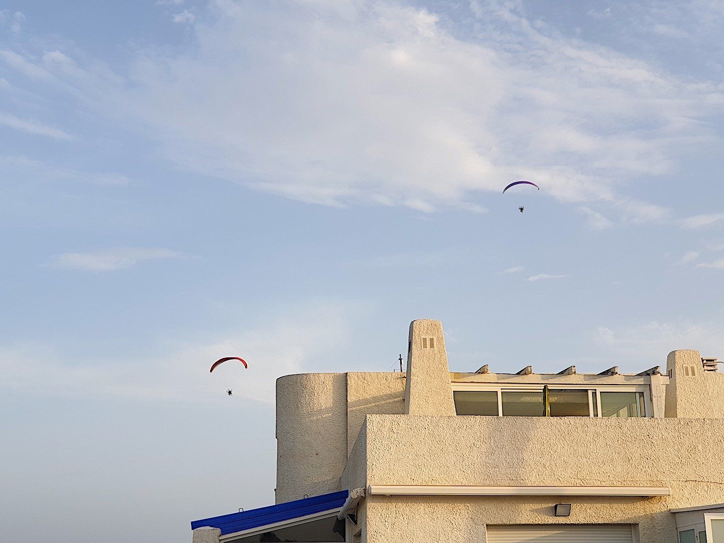 above the white building up in the blue sky are two paramotors flying high in the sky