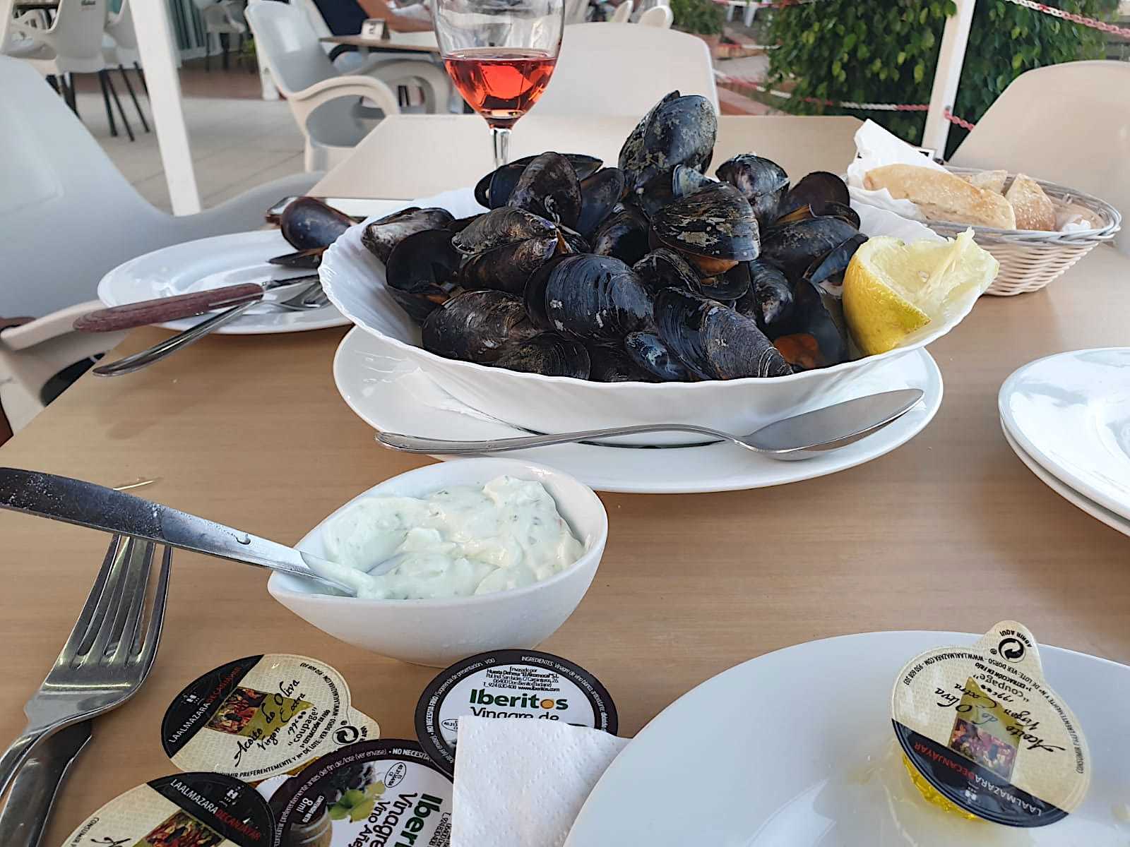 close up of bowl of oysters centre table with slice of lemon and some bread in a basket on one side and garlic butter in a dish on the other and a glass of rose wine in the background