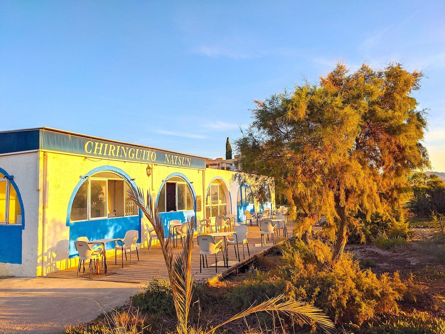 a white single storey building with big blue arches is lit up yellow by the morning sun