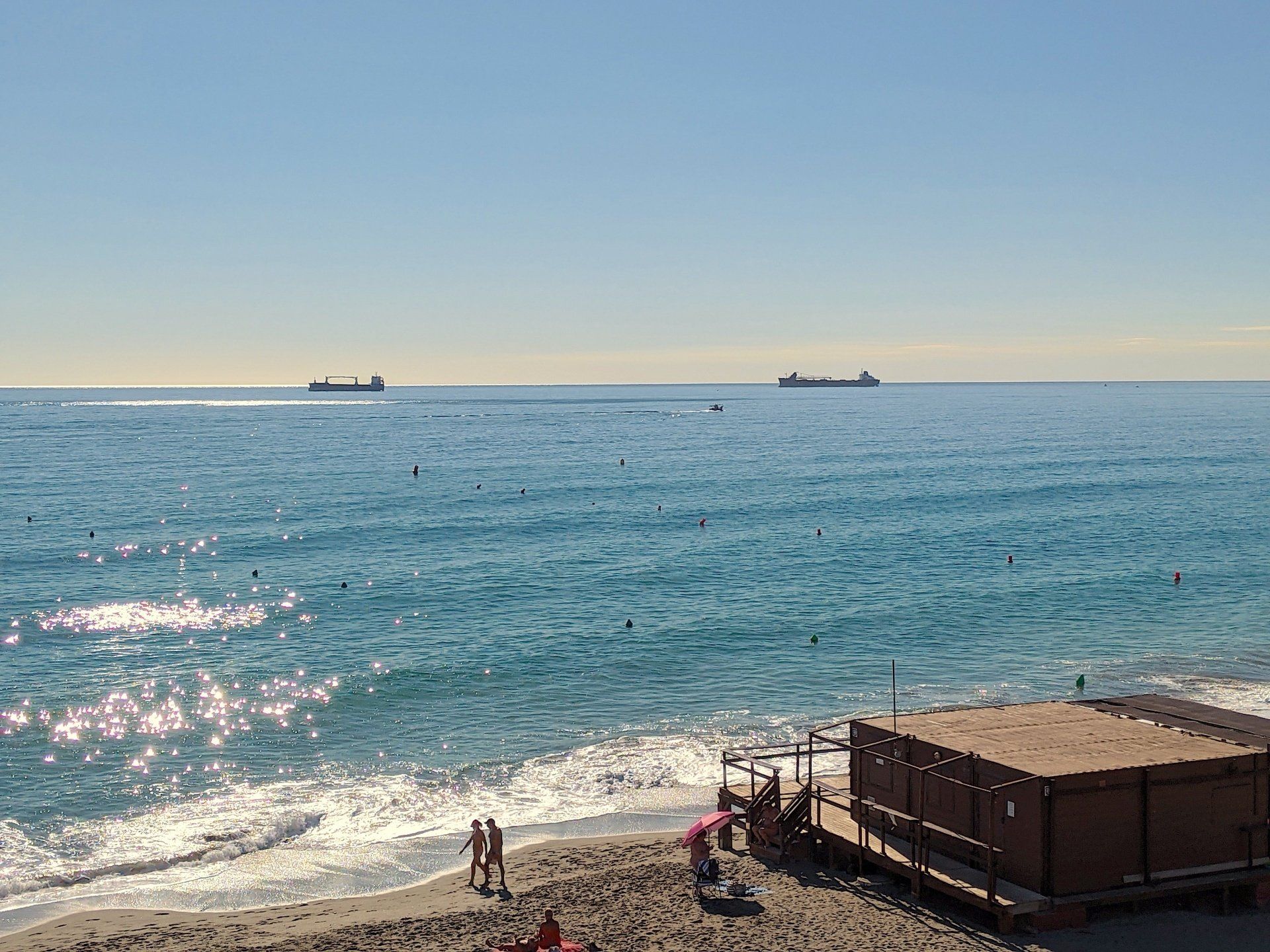 looking down onto a long sandy beach and a wooden beach bar at the waters edge and the water is blue and sparkling and there are two ships on the horizon