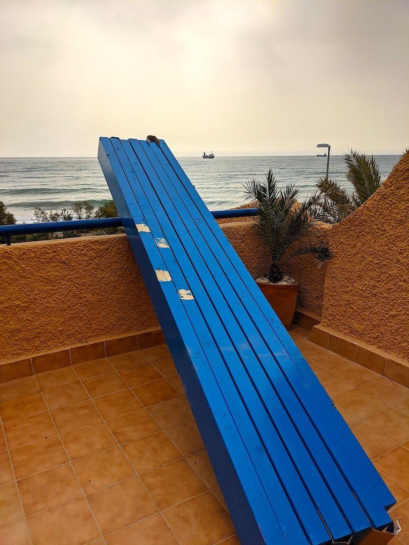 a pile of blue beams propped against an orange wall of a terrace with the sea in the background