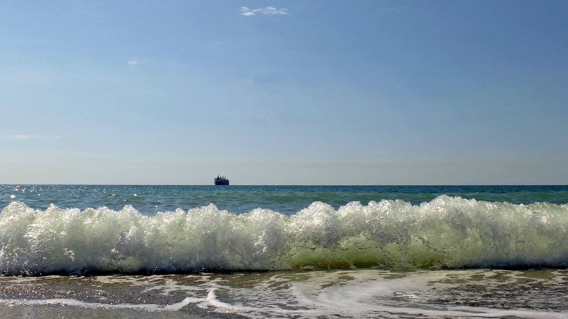 the rolling surf is coming towards you while beyond on the horizon there is a ship at sea