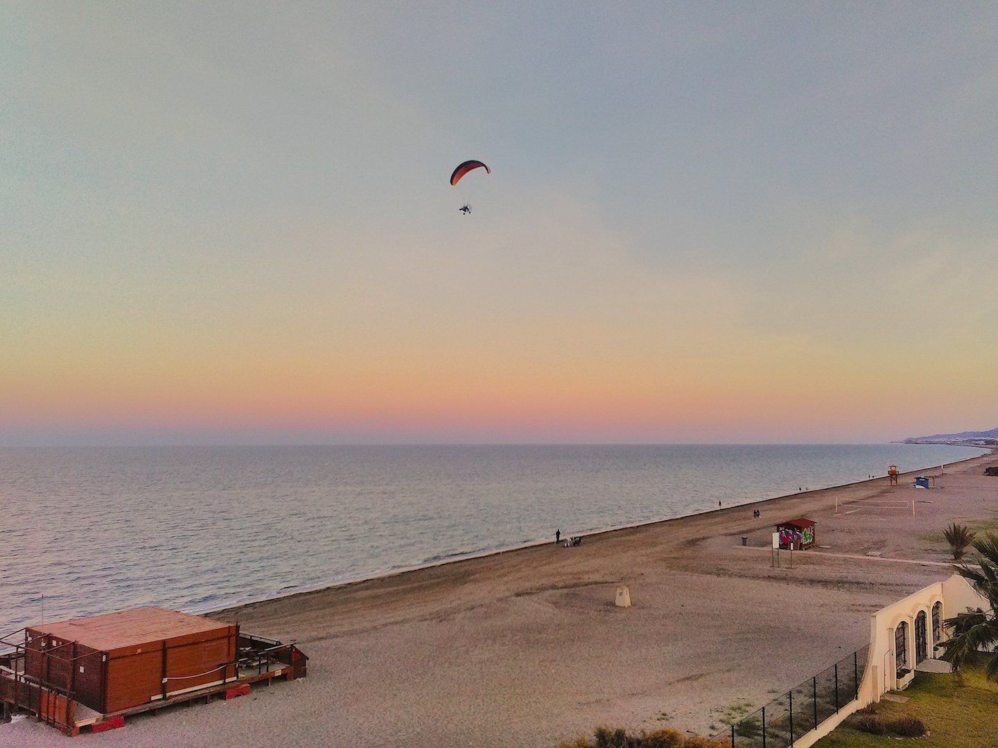 at sunset looking down the beach and a paramotor is flying high in the evening sky with colours of reds oranges and pinks on the horizon