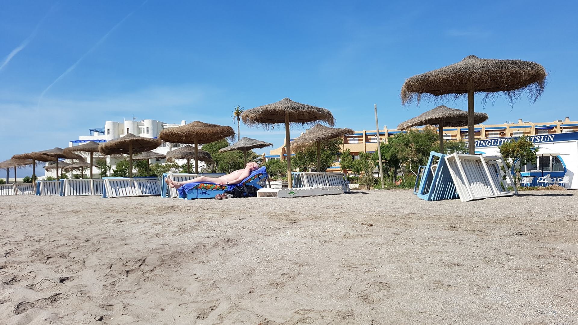 sun loungers with thatched parasols lined up on sandy beach with someone sunbathing naked on one of them