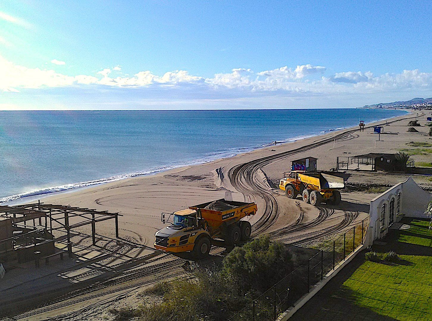 looking down from above onto the beach below and great big yellow dumper trucks are moving about the sand