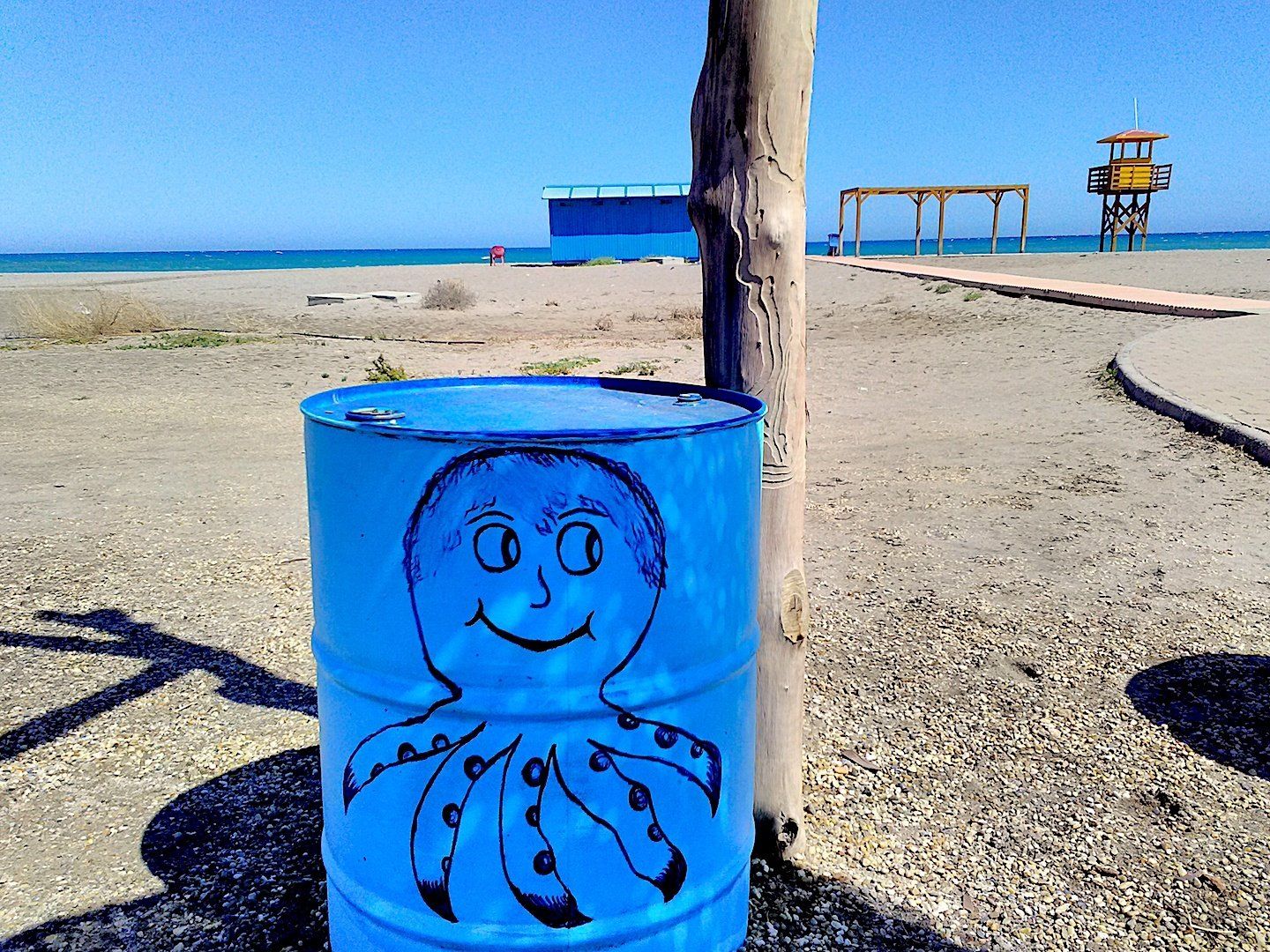 old oil barrel painted bright blue with an octopus painted on it and the sandy beach and sea behind