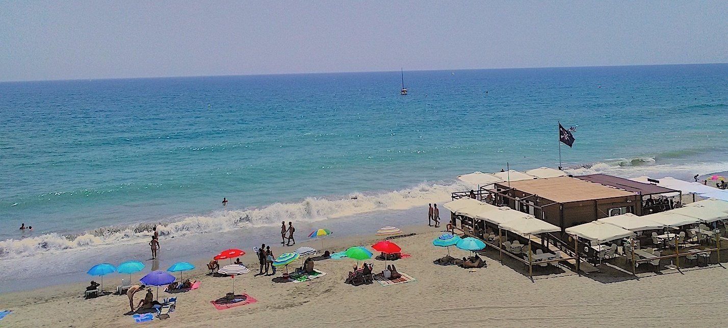 looking down onto warm looking beach and sea with sun brollies and wooden beach bar and people enjoying