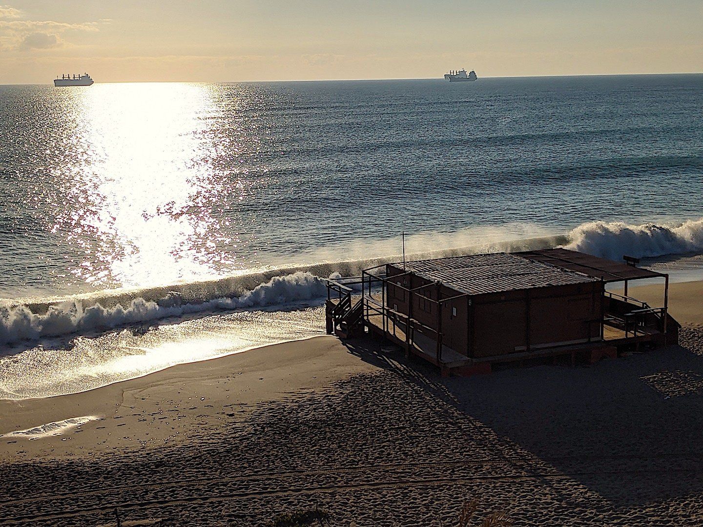 looking down onto the beach and the wooden beach bar at the shoreline where the surf is breaking at the waters edge