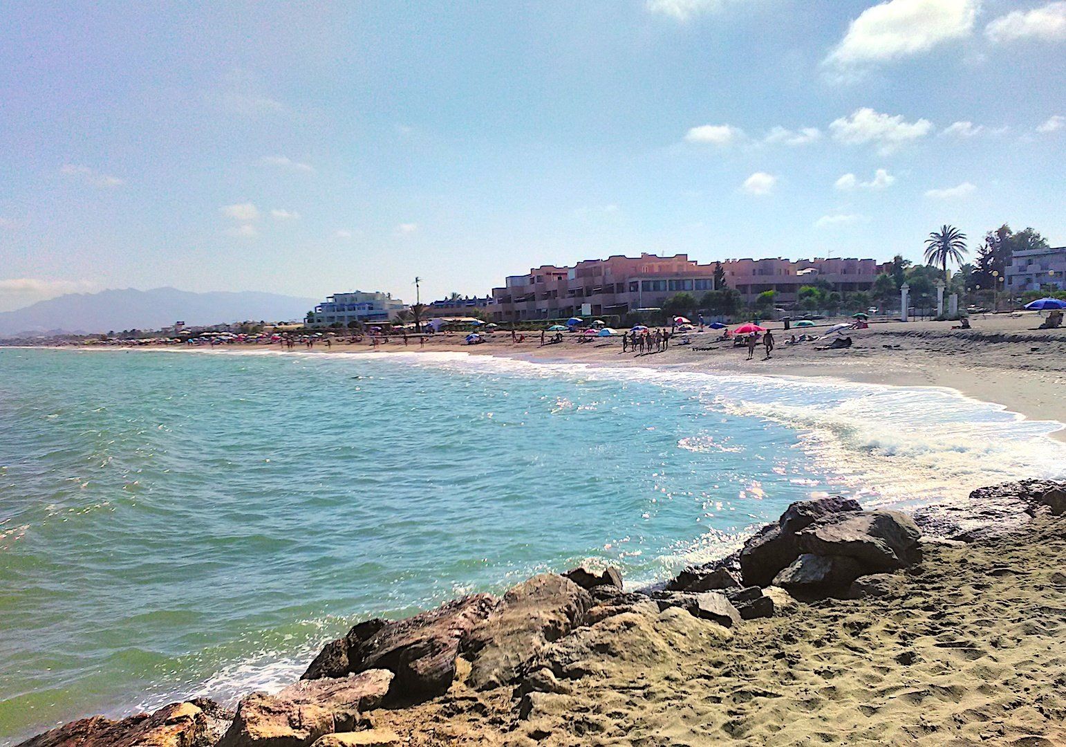 rocks with water splashing up in the foreground with the beach stretching away behind and lots of people on the beach