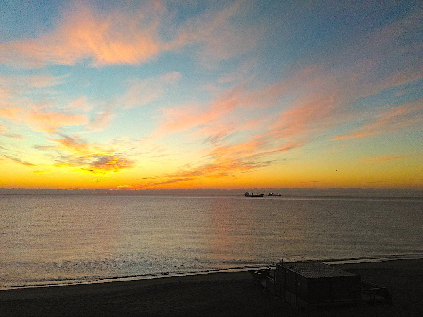 the beach at sunrise the horizon is yellow and gold and there are two big ships out at sea