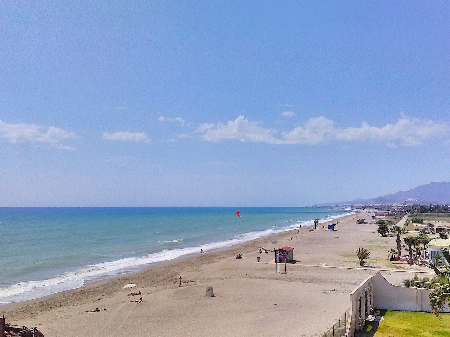 looking down the beach with a person hang gliding high in the blue sky