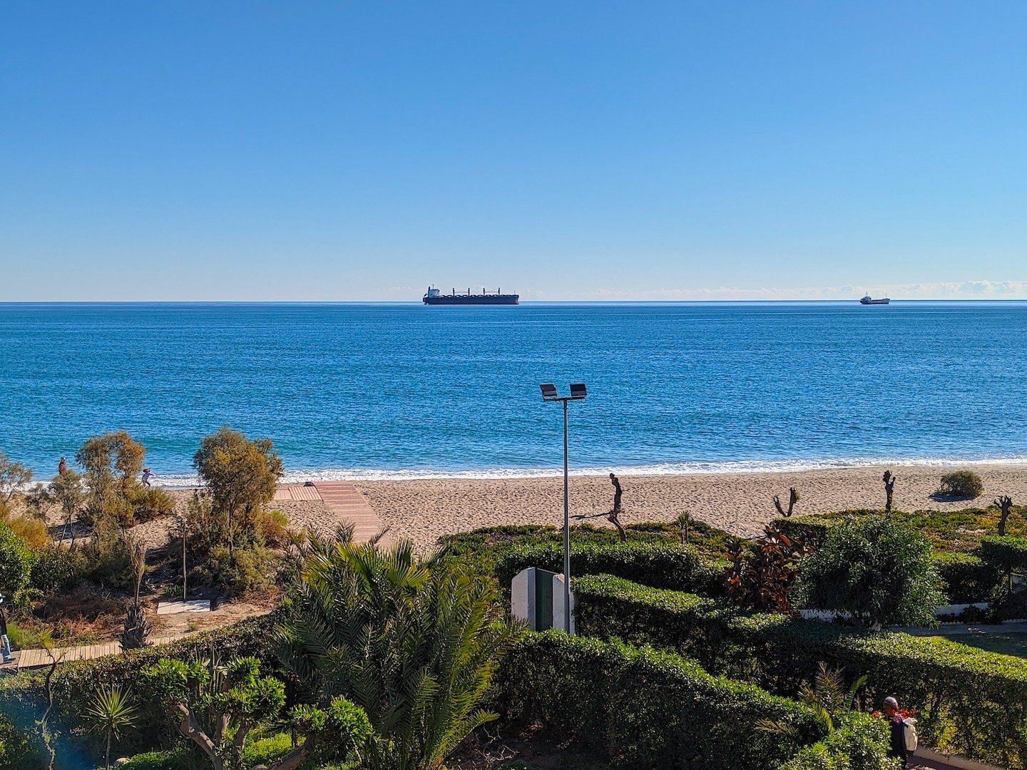 looking across gardens and hedges to a thin sandy beach and the sea beyond with two ships on the horizon