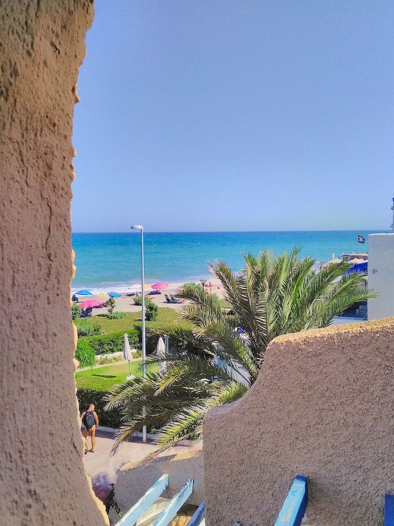 looking through a porthole on the terrace looking down onto footpath below and palm trees towards beach and blue sea in the distance