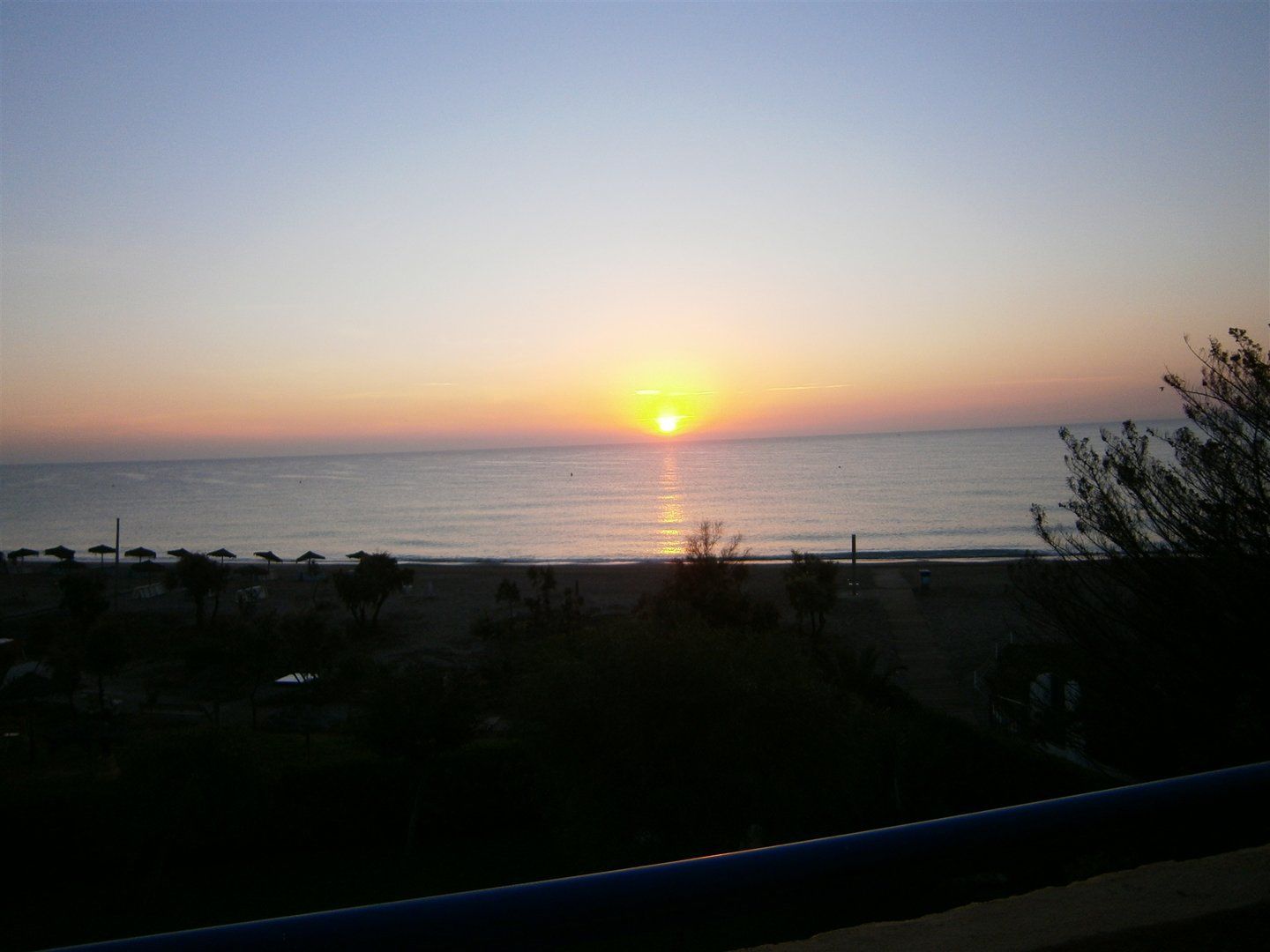 a beautiful yellow and orange sun is rising from the sea and you can see the thatched sun parasols on the beach