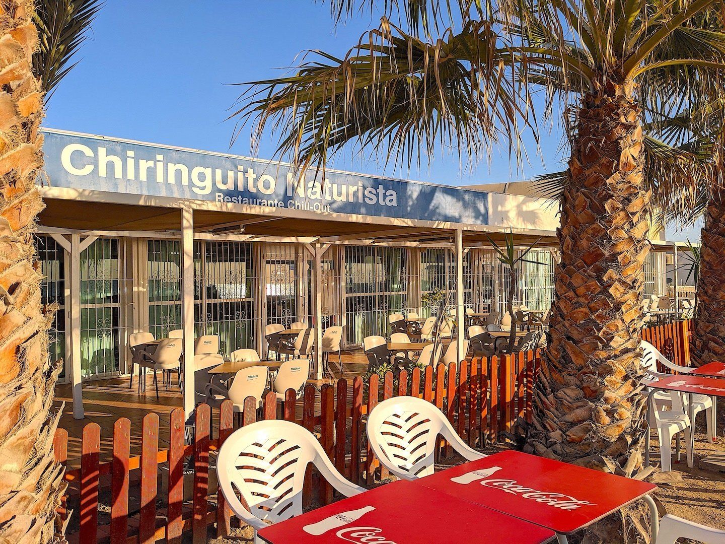two red tables side by side with white chairs and picket fence behind and restaurant building behind and palm tree to the right