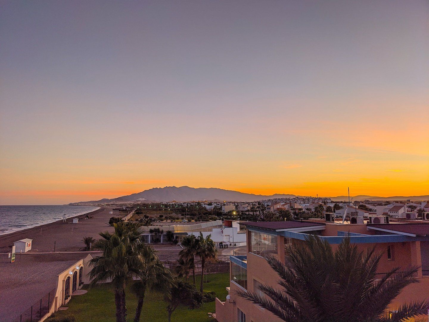 At sunset looking from the terrace across the tops of palm trees looking down the stretch of beach with the mountains in the distance all in orange and red colours