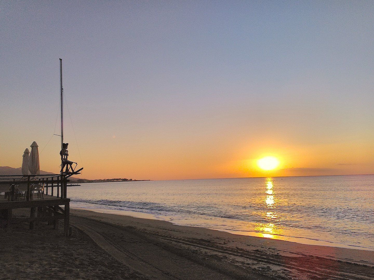 a skeleton on a pole looks out across the sea at dawn while the sun rises out of the horizon in the distance