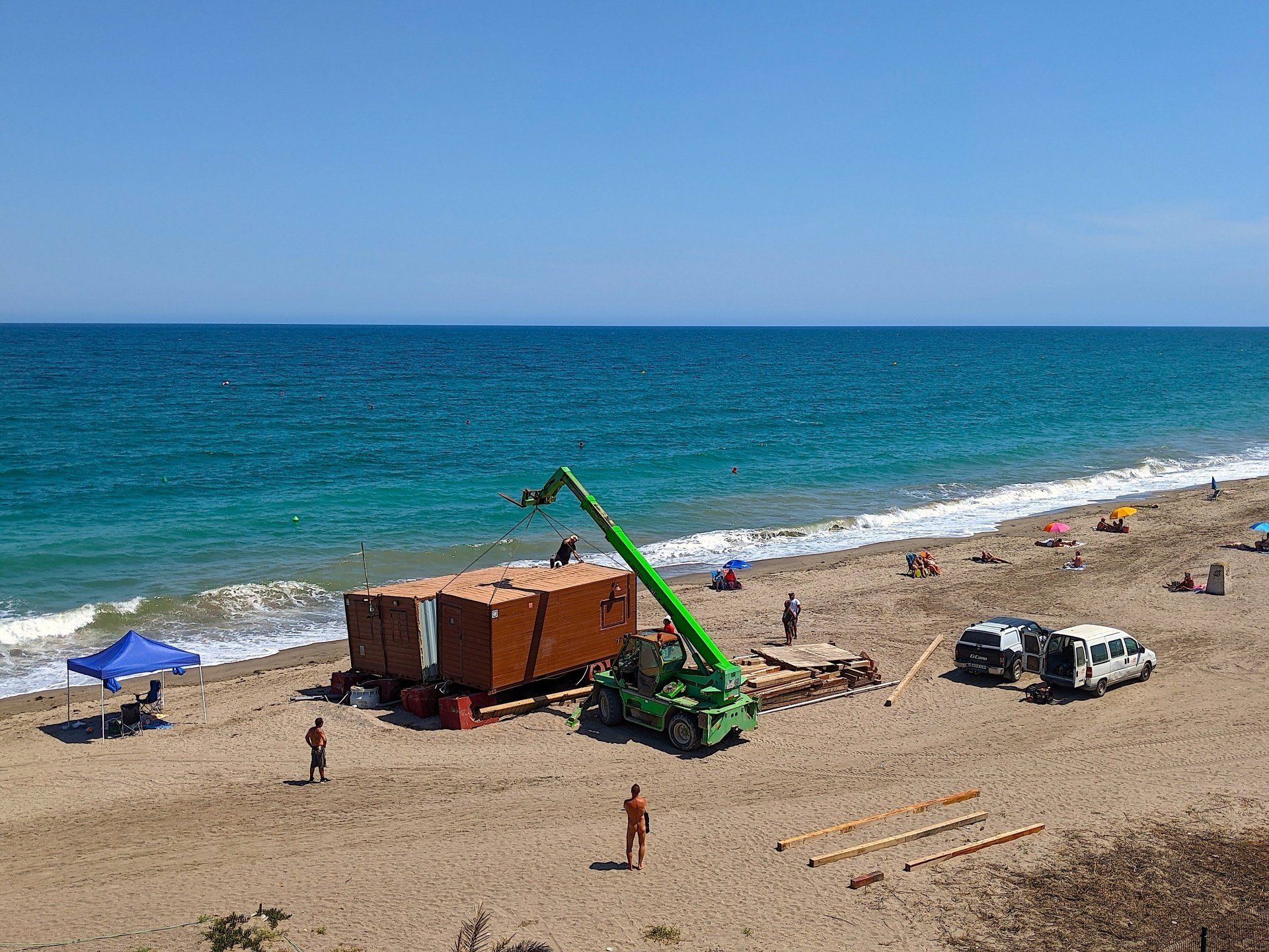 looking down onto the beach from above a crane is trying to lift the wooden beach bar and put it into a new position on the sand