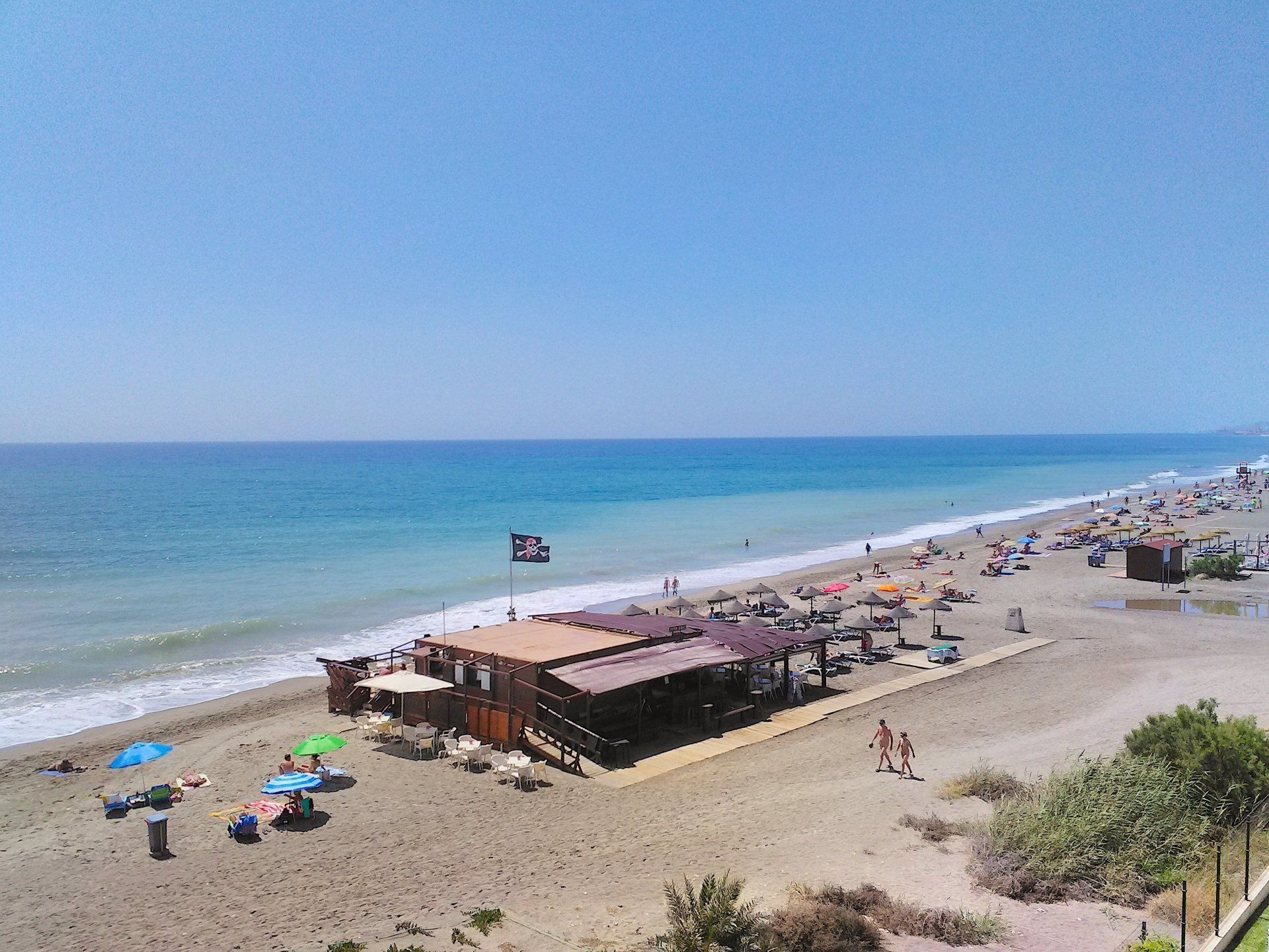 the sandy beach with lots of naked people enjoying the blue sky and warm sea with a wooden beach bar on the waters edge