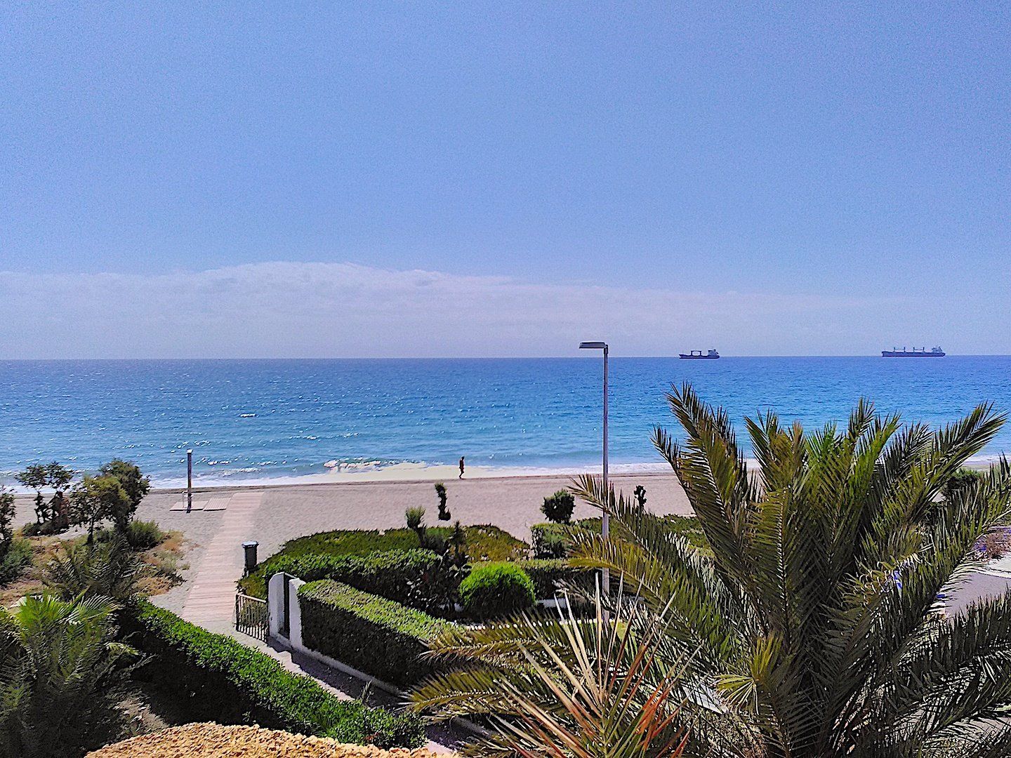 from the terrace looking over palm leaves onto a sandy beach below and blue skies and blue sea