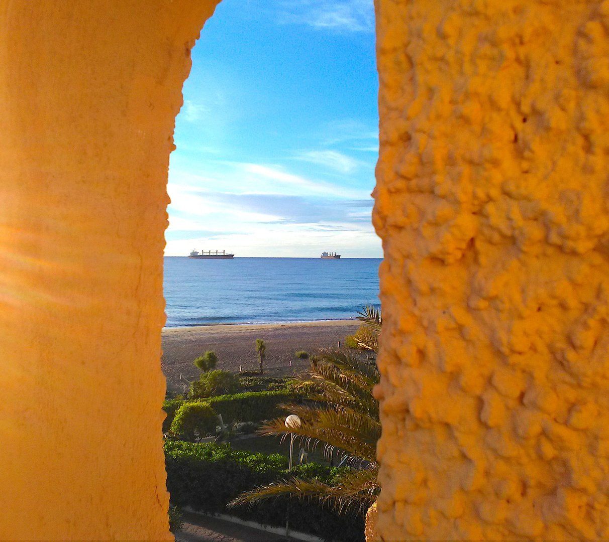 daytime looking through the porthole in the wall onto the palm leaves and two ships out at sea