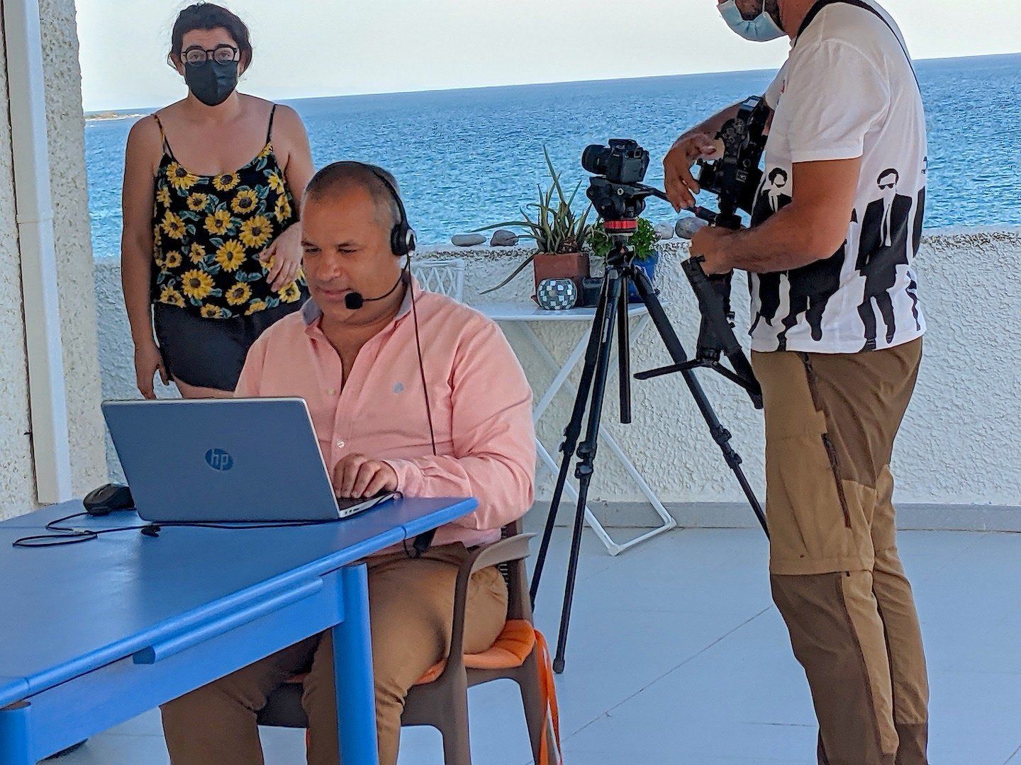man sat at table with a laptop working while two film crew people are behind him filming