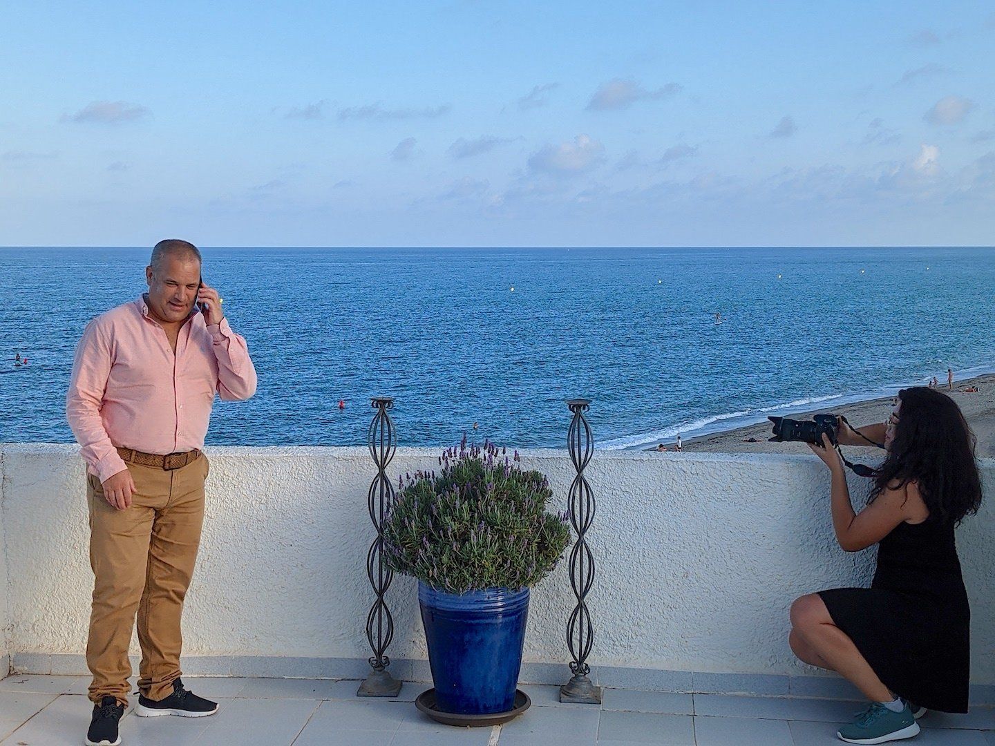 man stood talking on phone with sea behind him and woman with camera kneeling to take a photo