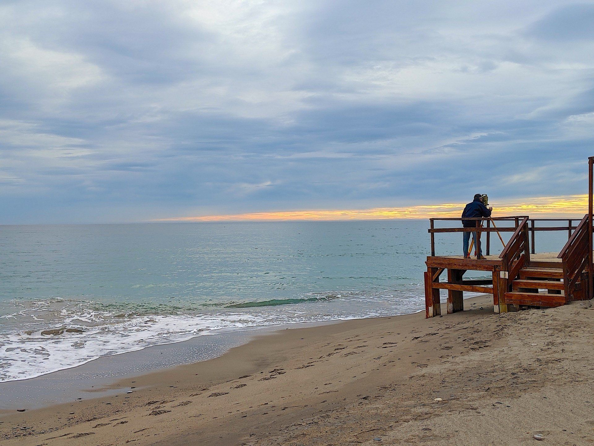 looking from the sand to a wooden shed on the beach at the waters edge there is a man with a special surveying tool on the decking