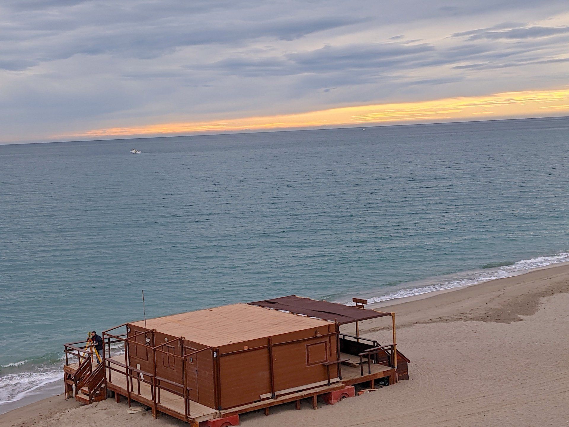 looking down from above onto a wooden shed on the beach at the waters edge there is a man with a special surveying tool in the bottom left