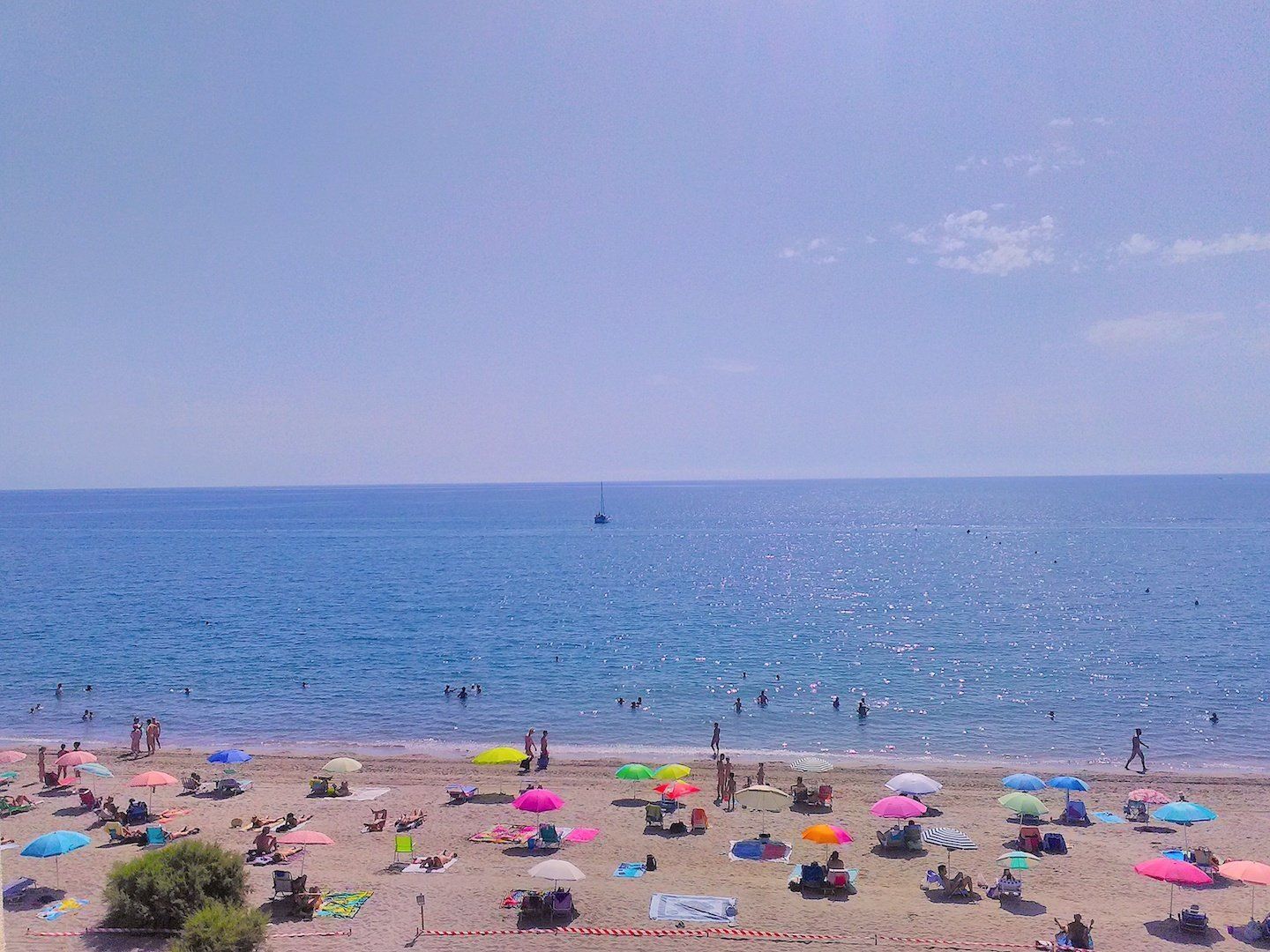 looking down onto a crowded sandy beach of multi colored sun umbrellas on a hot day with the sparkling blue sea beyond