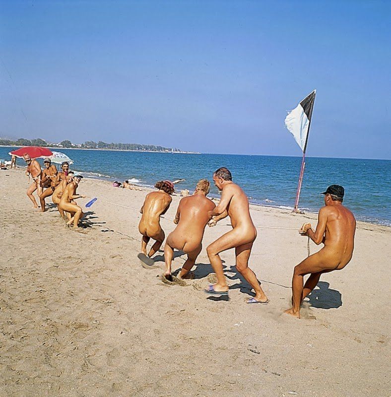 about ten people on a sandy beach on what looks like a hot day playing tug of war