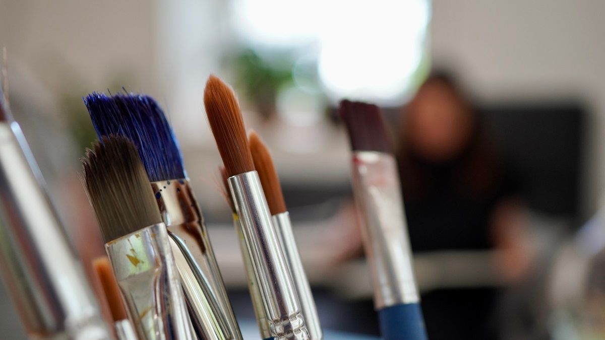 Verschiedene Pinsel im Vordergrund, im Hintergrund Susanne Grön in ihrer Praxis - Kunsttherapie Susanne Grön - Zarrentin