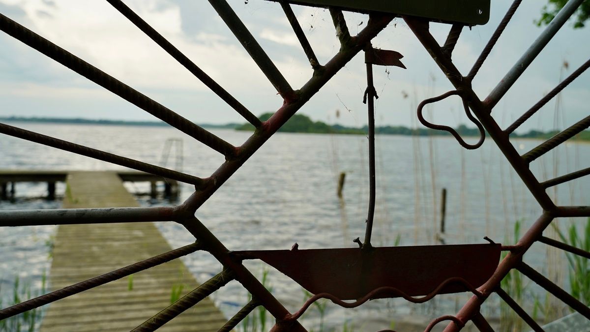 Blick durch einen Metallzaun hin zu einem See mit Steg - Praxis für Kunsttherapie Susanne Grön in Zarrentin.
