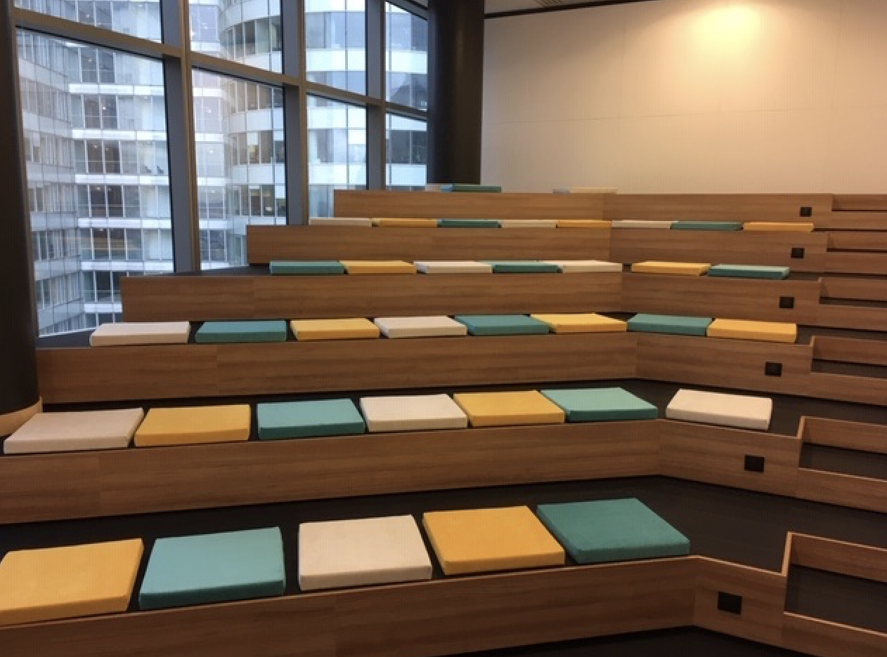 Gradins d'amphithéâtre en bois avec coussins d'assise rectangulaires colorés (bleu, vert, jaune, beige).
