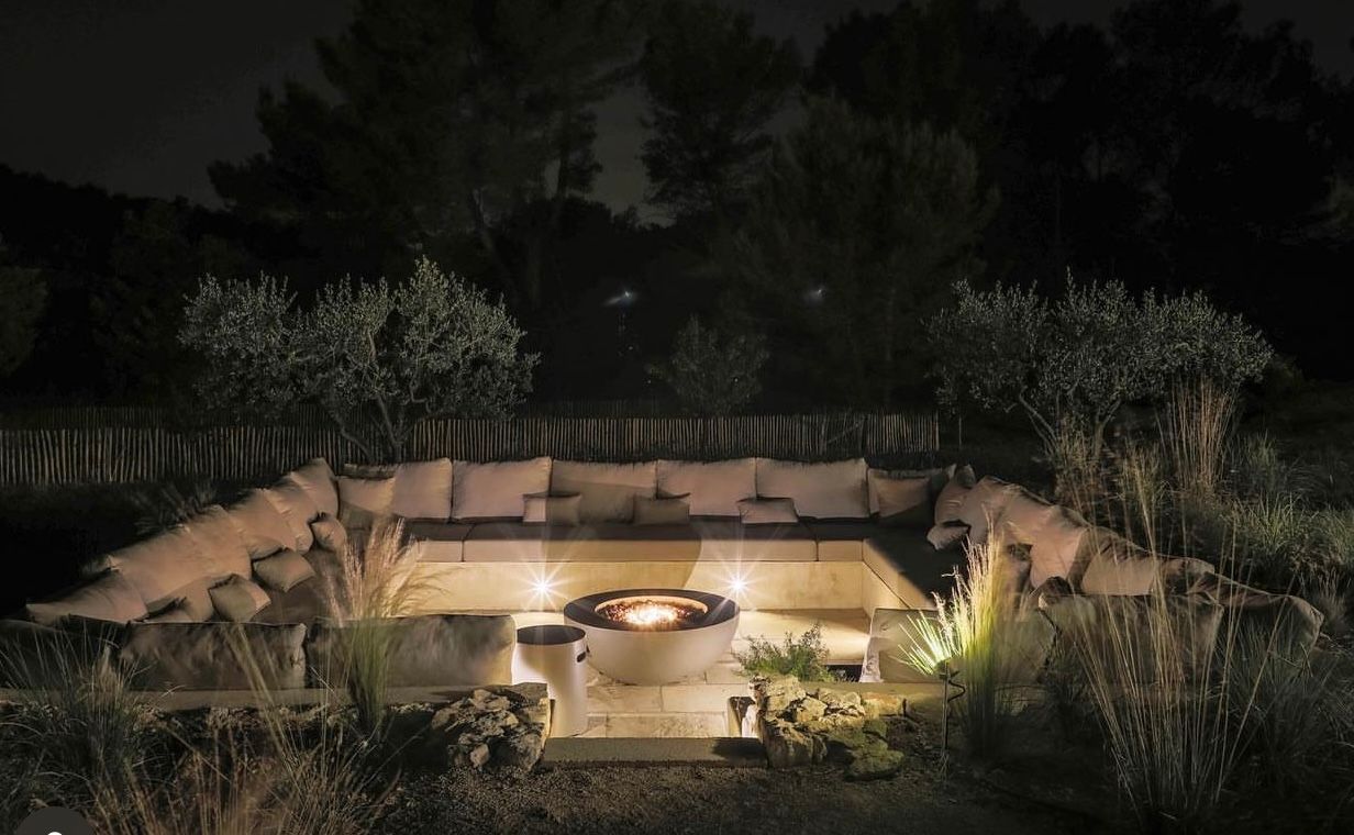 Espace lounge extérieur de nuit avec banquettes et coussins sur mesure autour d'un brasero éclairé.