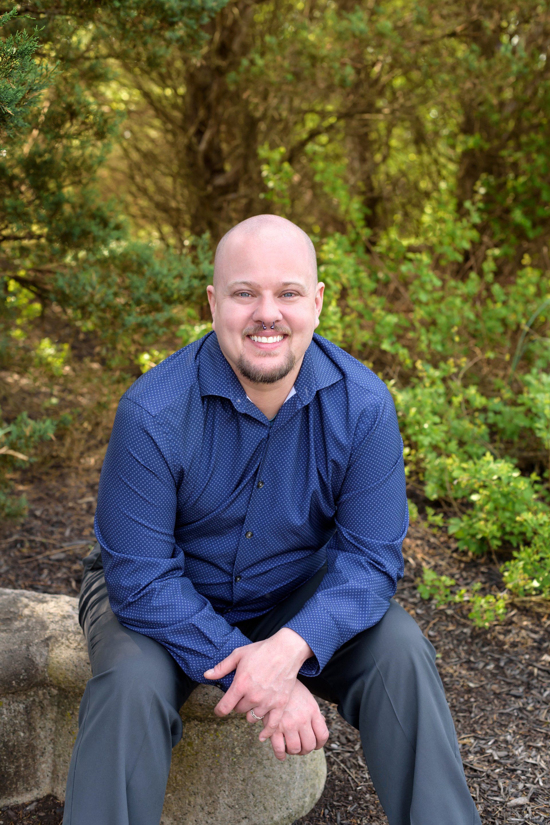 Daniel, a bald, smiling, white man wearing a blue shirt seated in a forested setting