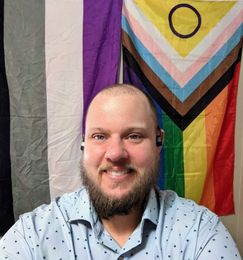 Nurse Daniel in front of a two flags: asexual pride and progress pride.
