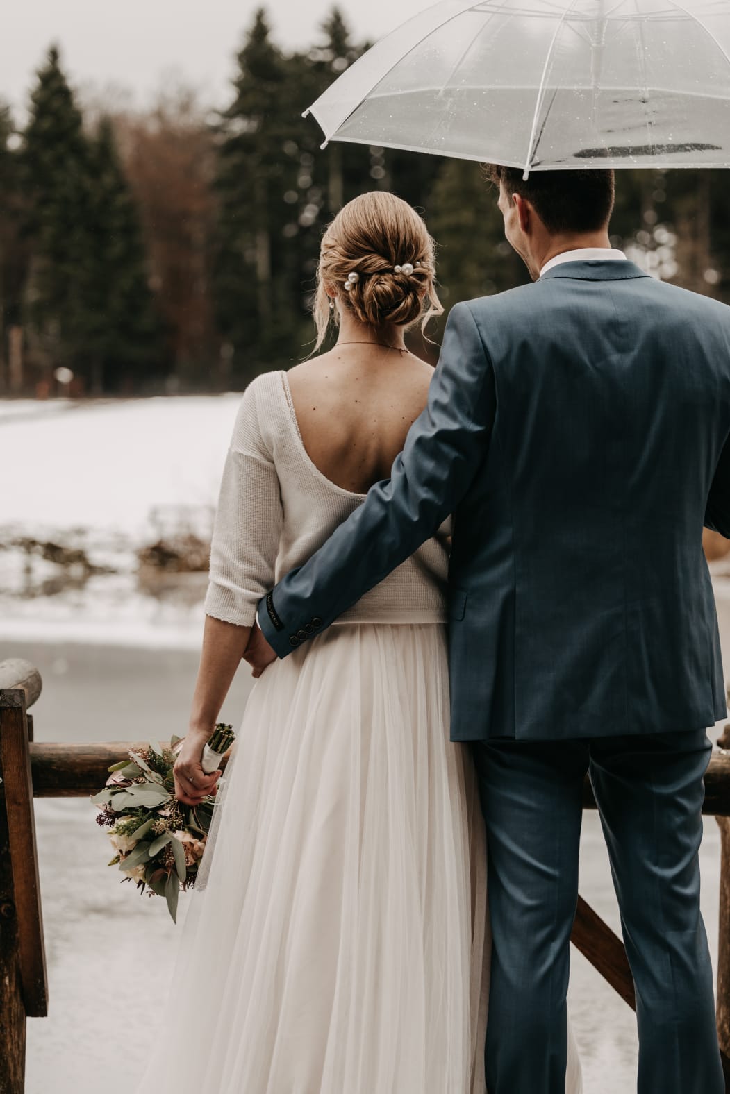 Braut und Bräutigam von hinten unter transparentem Regenschirm, eleganter Braut-Dutt und Hochzeitsstyling im Freien