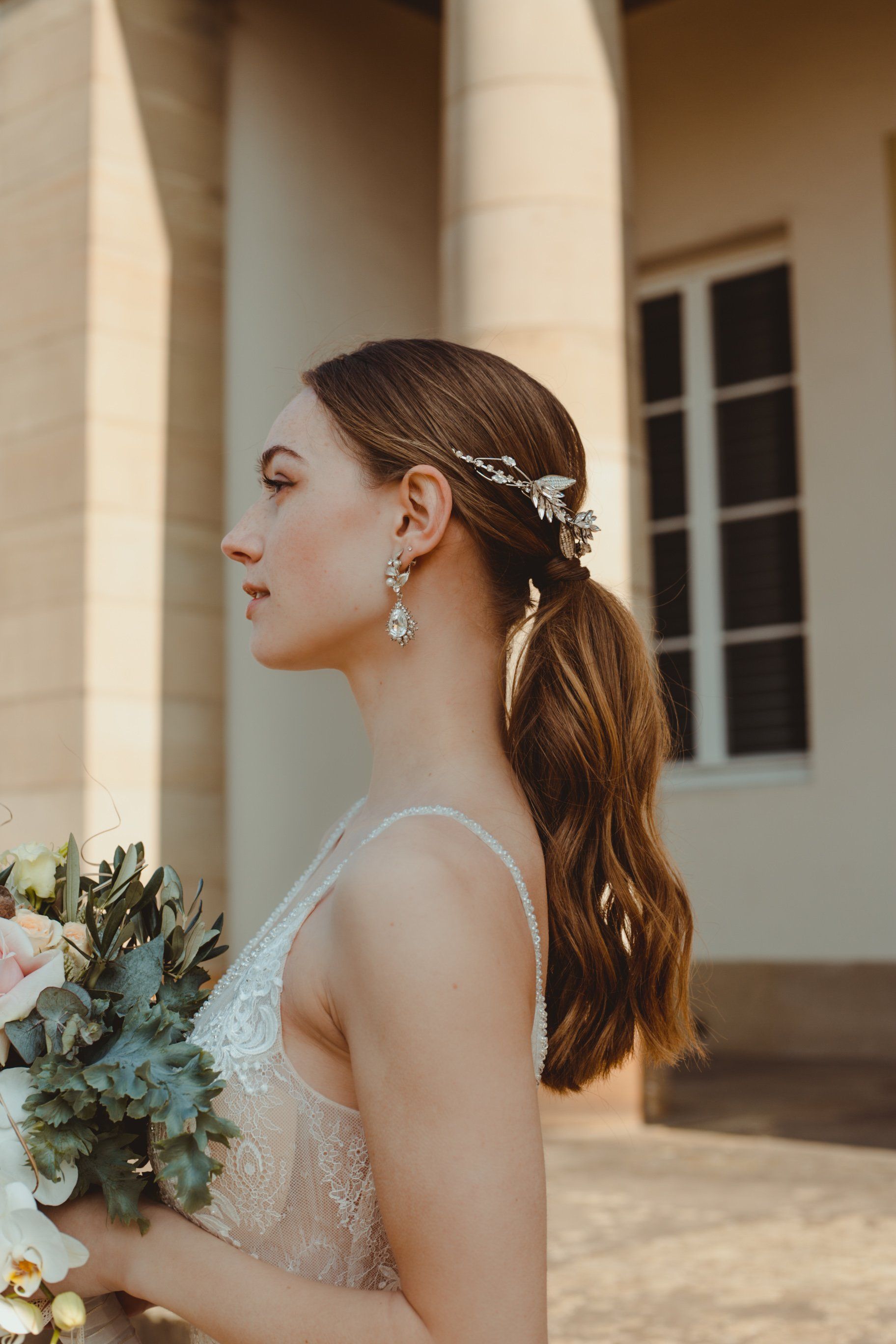 Braut im Profil vor Säulen eines schönen Gebäudes, glattes Brautstyling mit Haarschmuck und natürlichem Make-up