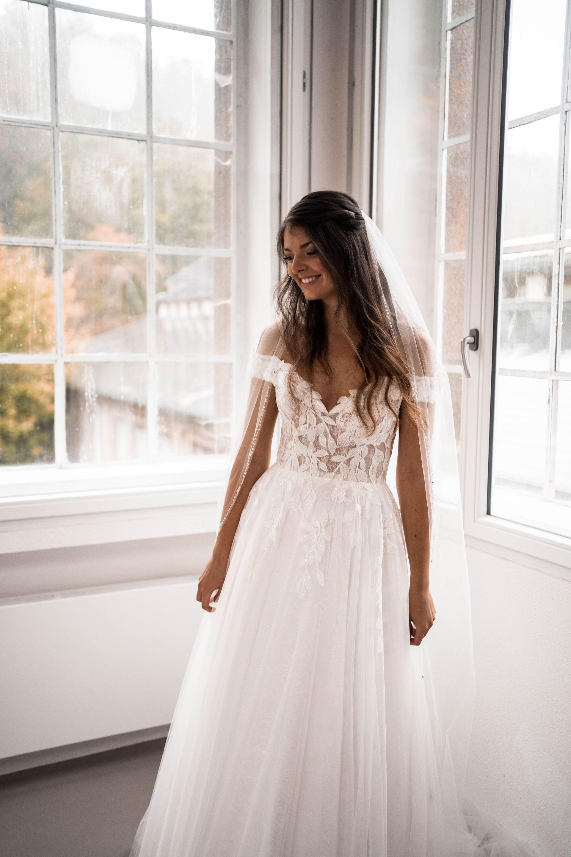 Braut lacht vor dem Fenster, natürlicher Hochzeitslook mit Schleier