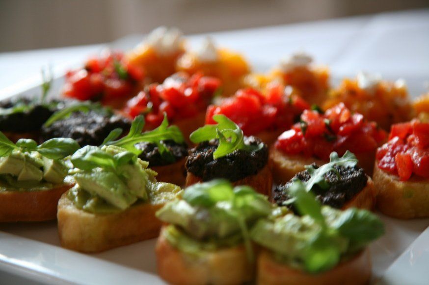 eine quadratische Platte mit Bruschette mit folgenden Belägen: Rucola-Parmesan, Tomate-Basilikum, Aprikose Ziegenkäse und Tapenade