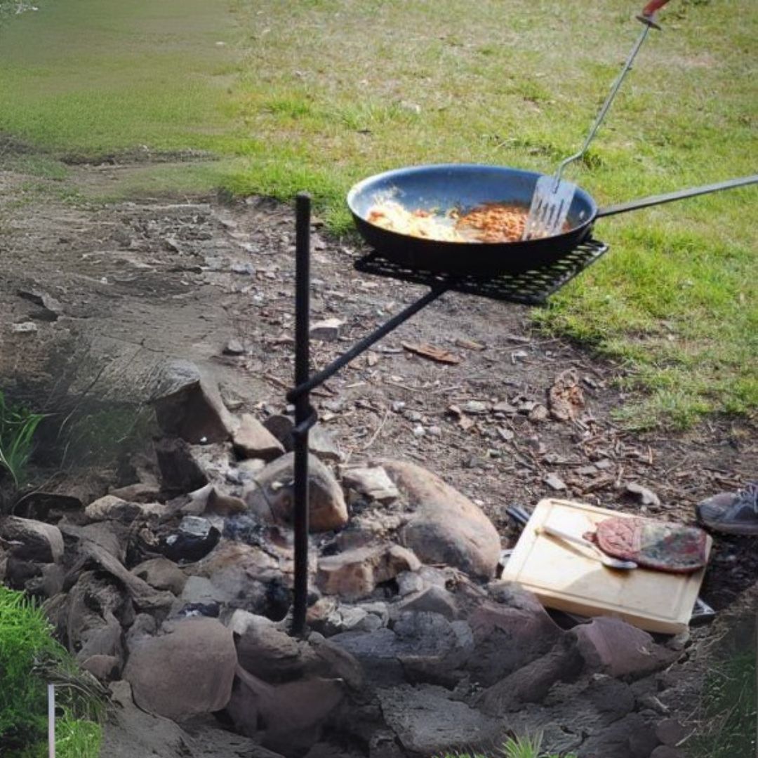 A simple black metal campfire cooking stake with a single attached grill grate holds a skillet filled with cooked food. A metal spatula rests in the skillet.
