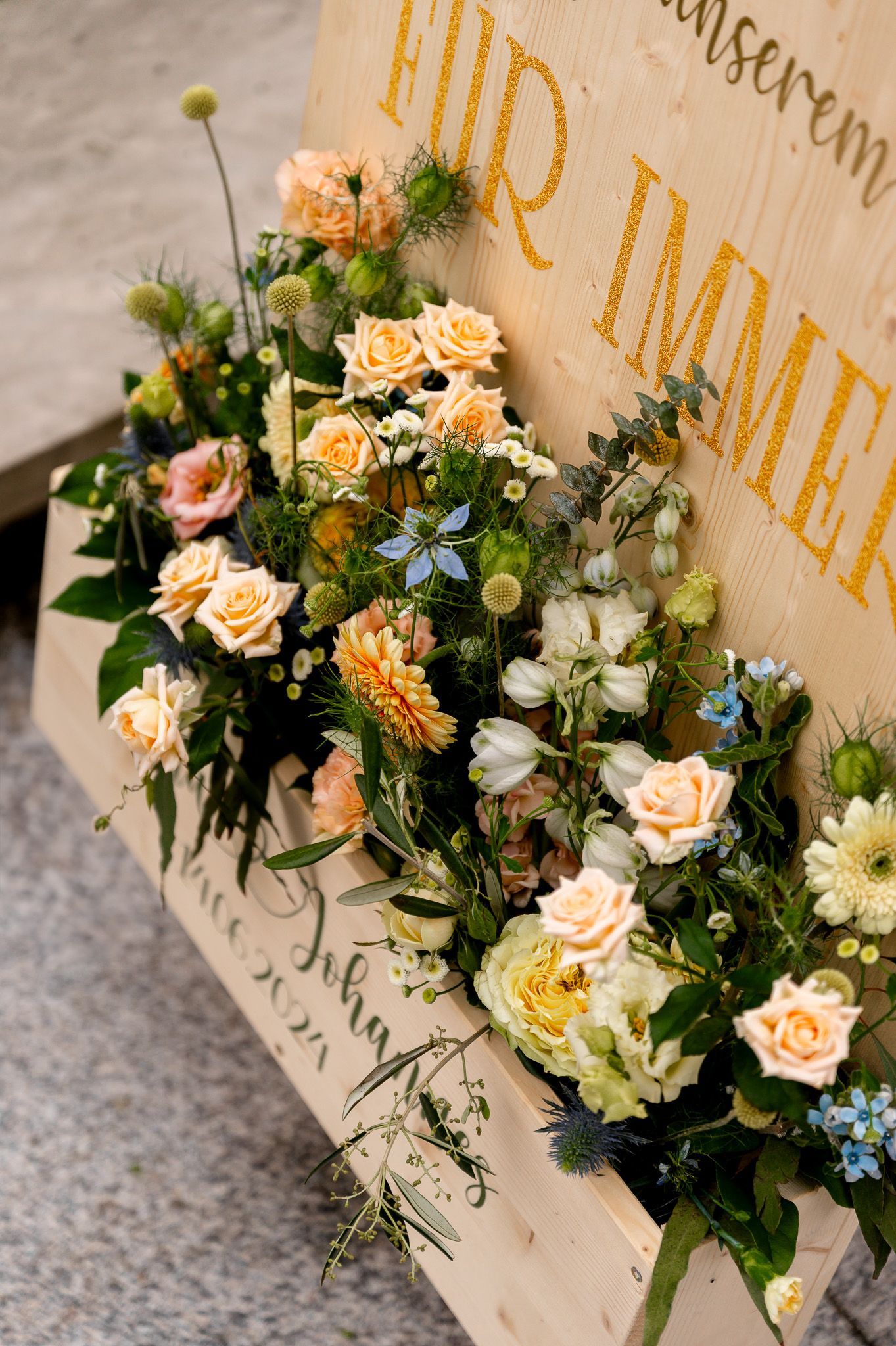 Detailaufnahme des Blumenschmucks am Hochzeitsschild mit cremefarbenen Rosen, Kamille, Eukalyptus und orangefarbener Gerbera.
