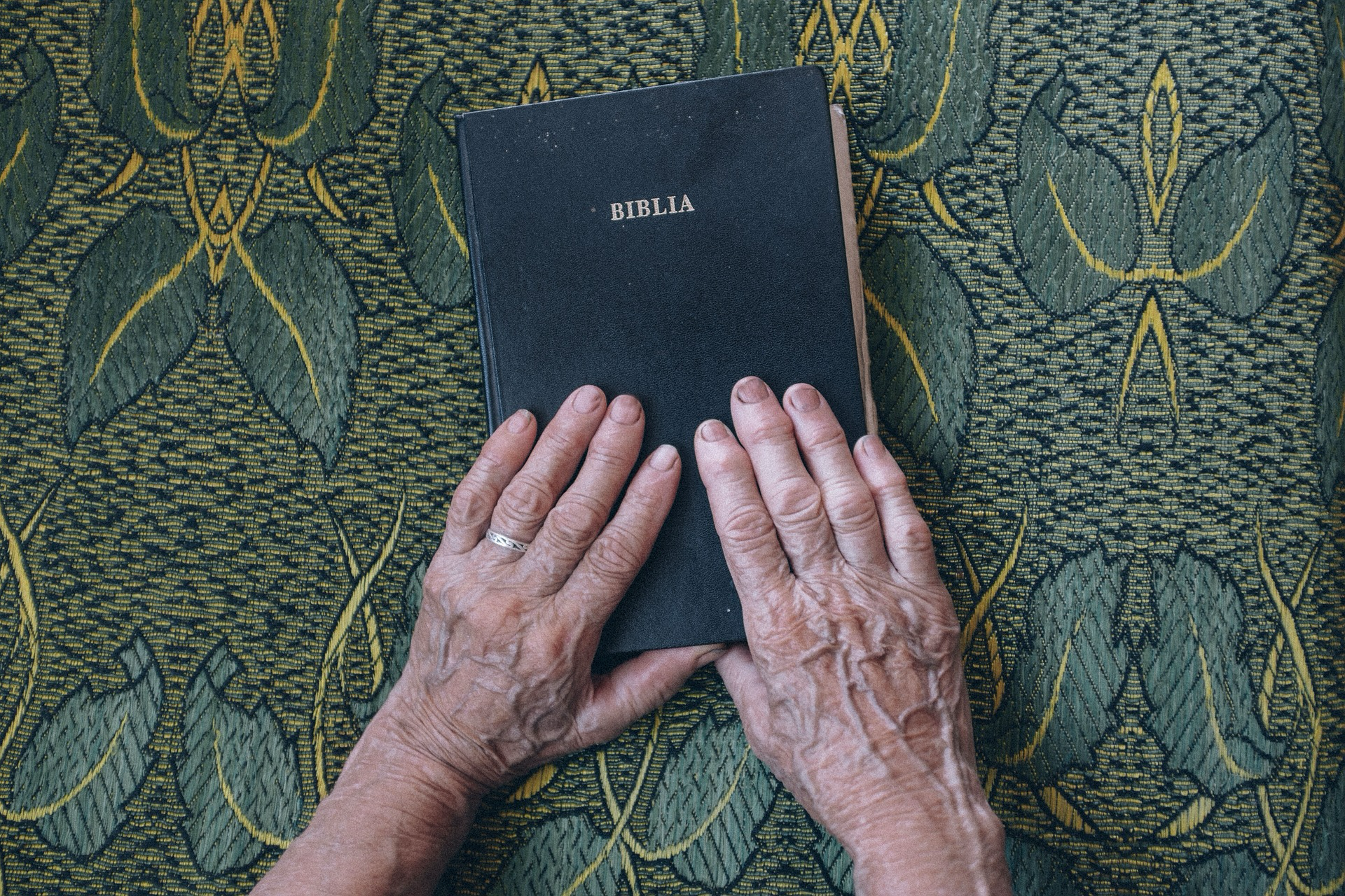 Auf dem Bild ist ein geschlossenes Buch mit dem Titel BIBLIA zu sehen. Es liegt auf einem Stoff mit einem Muster aus grünen Blättern und gelben Linien. Zwei Hände mit sichtbaren Alterszeichen und einem Ehering an der linken Hand ruhen auf dem Buch.