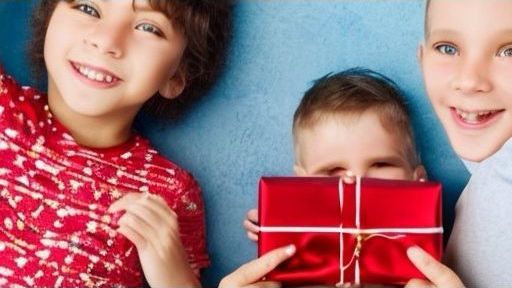 Auf dem Bild sind drei Kinder zu sehen, die auf einem blauen Hintergrund liegen. Das Kind in der Mitte hält ein rotes Geschenk vor sich. Das Kind links trägt einen roten Pullover mit einem Schneeflockenmuster und das Kind rechts hat ein hellblaues Oberteil an. Alle drei lächeln glücklich in die Kamera.