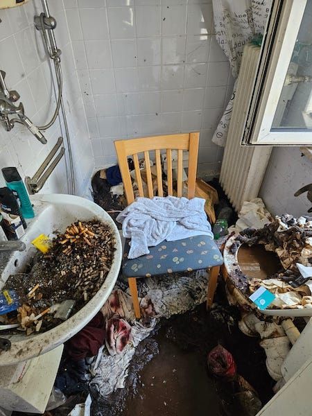 Das Badezimmer von einer Messi-wohnung. Alles ist dreckig. An den Wänden von der Dusche sind ganz viele Dreckflecken. Das Waschbecken ist voll mit zigarettenkippen. Der Boden ist total vermüllt und verdreckt.