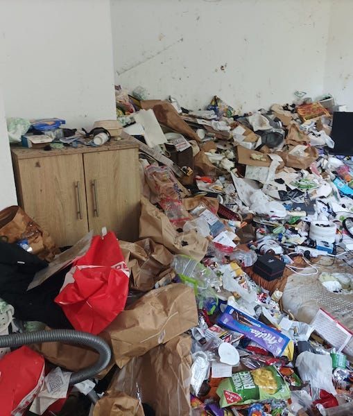 Ein mit Papiermüll überfülltes Messi wohnzimmer. An den Wänden haufenweise spinnenweben und schwarze striche. Alles ist dreckig.