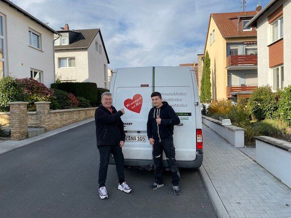 Entrümpler Gerald Molitor steht mit dem Mitarbeiter Raphael vor dem ENtrümpelungsauto und zeigt mit dem Finger auf das Logo: Entrümpelung mit Herz.